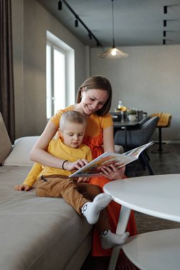 Kanepede oturup resim kitabı okuyan gülümseyen anne ve çocuğun dikey portresi.
