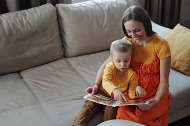 Masal kitabındaki resimlere bakarak gülümseyen anne kitap okurken kanepenin üzerinde birlikte oturup boşluğu kopyalarken yüksek açılı görüntü.