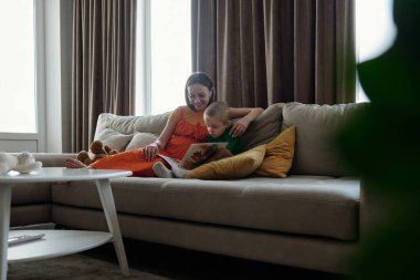 Geniş açılı çekimde, genç oğluyla kitap okuyan hamile bir kadın ön planda yeşil bitki çerçeveli bir kanepede otururken.