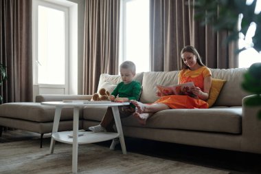 Genç çocuğun kille oynadığı ve hamile annenin kanepe yeşili çerçevesinde rahatlarken kitap okuduğu uzun bir çekim.