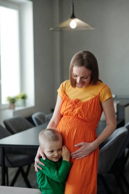 Küçük oğlu olan hamile bir kadının dikey portresi. Mutfakta durmuş karnını dinliyordu.