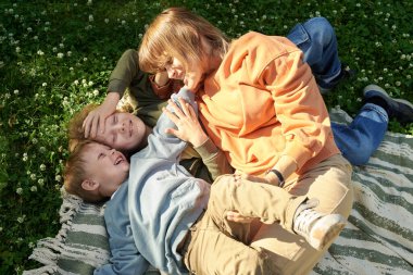 Parktaki yeşil çimlerin üzerinde piknik battaniyesinde uzanan iki oğlana sarılan gülümseyen genç annenin yüksek açılı görüntüsü