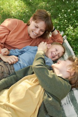 İki küçük kardeşin dikey portresi, yeşil çimlerin üzerinde annesiyle piknik battaniyesinde yatarken eğleniyor ve gülümsüyor.