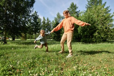 Annesinin küçük çocukla el ele tutuşup açık havada güneşli parkta yeşil çimlerde oynadığı geniş açı görüntüsü.