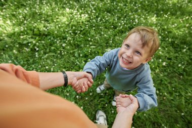 Yeşil parkta el ele tutuşup oynarken annesine bakan gülümseyen genç çocuğun samimi yakın çekimi.