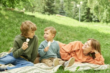 Anne portresi ve iki küçük çocuk parkta eğleniyorlar ve piknik battaniyesinde oturmuş dondurma yiyorlar.