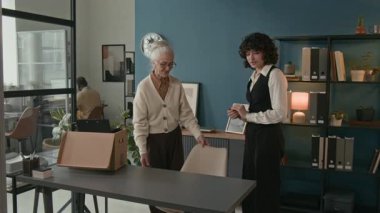 Orta yaşlı, beyaz bir kadının, emeklilik günü için modern ofiste genç yerine geçecek kişiyi karşılarken ve brifing verirken neşeli bir şekilde ofis eşyalarını teslim ettiği bir fotoğraf.