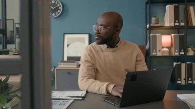 Belinde bej renkli süveter giyen genç bir adam düzenli masasında çalışıyor. Etrafı dokümanlar ve modern ofis içi ile çevrili. Verimlilik ve konsantrasyonu yansıtıyor.