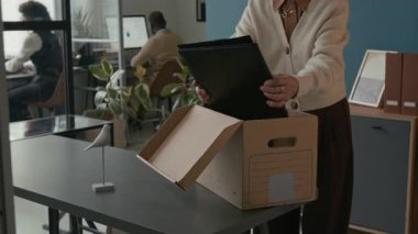 Kafkasyalı üst düzey bir kadının ofis emekliliği sırasında çalışma alanını toparlarken arka planda çalışan meslektaşlarının evrak ve klasörleri karton kutuya yerleştirirken çekilmiş görüntüleri.