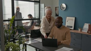 Üst düzey beyaz kadın, emekliliğinden önce dizüstü bilgisayarda oturan genç Afrikalı Amerikalı meslektaşına arka planda çalışan diğer takım üyelerine rehberlik sunuyor.