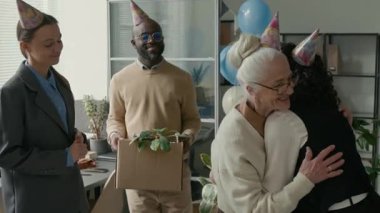 Yaşlı emekli, son gününü parti şapkaları ve balonlarla kutlarken genç meslektaşlarıyla sıcak bir kucaklaşma paylaşıyor. Afrikalı Amerikalı erkek iş arkadaşı ona karton kutu dolusu eşyayla yardım ediyor.