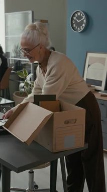 Son gününde parlak, rahat ofis ortamında ofis eşyaları paketlerken gülümseyen yaşlı beyaz kadının dikey orta pozu.
