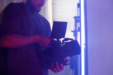 Prodüksiyon ekibinin elinde profesyonel video kamera tutması ve stüdyo çekimi sırasında kontrolleri ayarlaması.