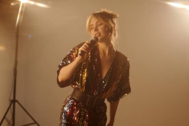 Klip çekimlerinde mikrofona şarkı söyleyen genç bir kadının belden yukarı portresi.