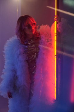Genç bir kadının dikey portresi. Stüdyoda çekimler sırasında farklı renklerde neon ışıklı şeritte duruyor.