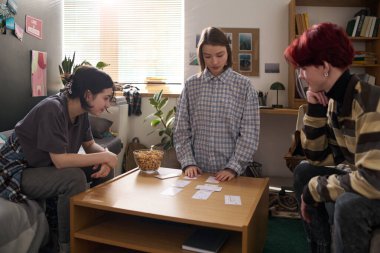 Evde ya da yurt odasında birlikte kağıt oynayan üç genç kız grubu. Masanın üzerine kart seren kız odaklı.