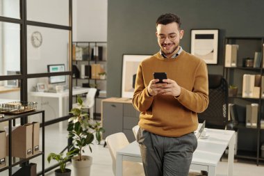 Modern ofis ortamında durup sosyal medyayı kontrol ederken akıllı telefon kullanan Orta Doğulu bir gencin portresi, boşluğu kopyala