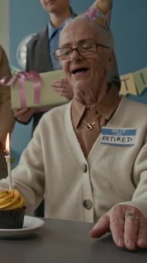 Mutlu ve heyecanlı yaşlı bir kadın emeklilik partisi sırasında kekin üzerine mum üflüyor. Etrafı neşeli iş arkadaşlarıyla çevrili ve güzel süslenmiş ofis hediyeleriyle dolu.