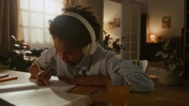 Kulaklıkları takılı ve elinde kalemle, Afrikalı Amerikalı okul çocuğu ev ödevlerine konsantre oluyor, rahat, bitki dolu bir odada.