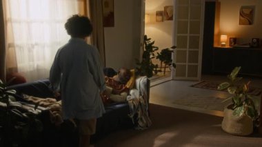 Yumuşak ışık ve sıcaklıkla dolu sessiz bir evde çocuk, dinlenen babasının üzerine düşünceli bir battaniye örterek günlük şefkat anı yaratıyor.