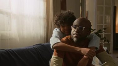 Afro-Amerikan baba ve oğul arasındaki sıcacık kucaklaşmayı yakalayan neşeli orta boy portre, her ikisi de sıcak oturma odalarına akan güneş ışığı gibi ışıl ışıl gülüyor.