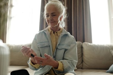 Son sınıf öğrencisi kanepede oturup sıcak, rahat bir evde glukometre kullanarak kan şekerini ölçüyor. Arka pencereden odayı doğal ışık kaplıyor.