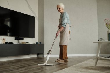 Üst düzey kadın oturma odasının zeminini süpürmek için süpürge kullanıyor. Sıradan giyinmiş, memnun görünüyor ve yaşlılar için aktif yaşam tarzını teşvik ederek görevine odaklanmış görünüyor.
