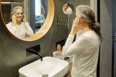Modern banyoda duran yaşlı bir kadın, beyaz bluz giyerken aynaya gözlük takıyor. Banyo lavabo, musluk ve yansıtıcı yüzey içerir.