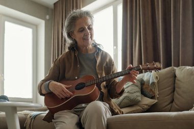 Son sınıf öğrencisi bir kadın rahat bir koltukta oturup gitar tıngırdatıyor ve hafifçe gülümsüyor. Geniş pencerelerden sızan doğal ışık odası sıcak ve davetkar bir atmosfer yaratıyor.