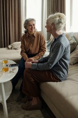 Oturma odasındaki kanepede oturan iki yaşlı kadın pencereden içeri güneş ışığı giriyor ve çay fincanlarıyla gülüyor ve kaynaşıyorlar.