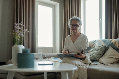 Kanepede oturan yaşlı kadın elinde defterle oturma odası düzeninde notlar yazıyor. Aktivite sırasında gözlük takıyor ve rahat kıyafetler giyiyor.