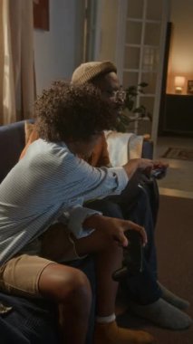 Dikey çekimde, Afrikalı Amerikalı baba ve oğul, odaklanmış ve heyecanla dolu, yan yana oturuyor, kontrolörleri kavrıyor ve sıcak ışıklı oturma odalarında oyun oynarken kahkahalarını paylaşıyorlar.