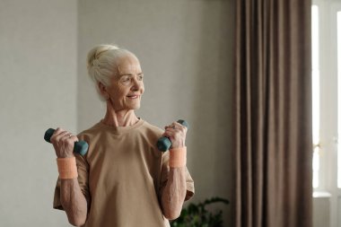 Yaşlı bir kadın kollarında halterlerle egzersiz yapıyor, tarafsız renkli gömlek giyiyor, pencerenin yanında duruyor, perdelerle örtülü, gülümsüyor ve yan tarafa bakıyor.