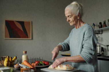 Mutfak tezgahında dikilmiş taze sebzeler ve malzemelerle ekmek dilimleyen kıdemli bir kadın. Arka planda görünen modern mutfak dekoru