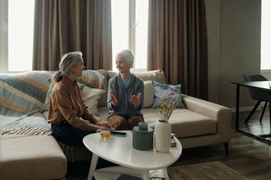 Son sınıf kadınları kanepede oturuyor, canlı sohbetler yapıyorlar ve rahat bir oturma odasında doğal ışıklandırma ve rahat bir ortam ile hikayeler paylaşıyorlar.