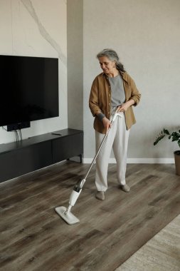 Yaşlı bir kadın modern oturma odasında ahşap zemini buharlı paspas ile temizliyor. Arka planda minimalist dekor ve duvar destekli TV standı var. Yanında da yeşillik var.