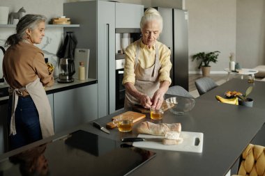 Üst düzey bir kadın mutfak tezgahında malzemeleri hazırlarken genç bir kadın bakıyor, ikisi de önlük giyiyor ve mutfak aletleri tezgahta.