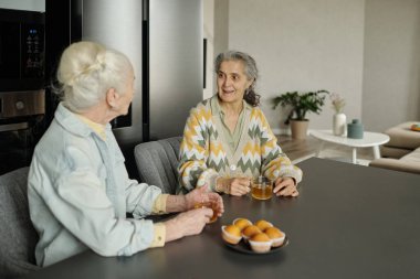 Mutfak masasında içkilerle ve yeni pişmiş çöreklerle oturup sıcak ve misafirperver bir ortam yaratarak sohbet eden iki son sınıf öğrencisi kadın.