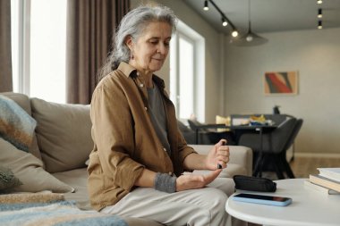Yaşlı bir kadın kanepede oturuyor ve rahat oturma odasındaki aletleri kullanıyor. Akıllı telefonu ve diğer cihazları masaya yerleştiriyor.