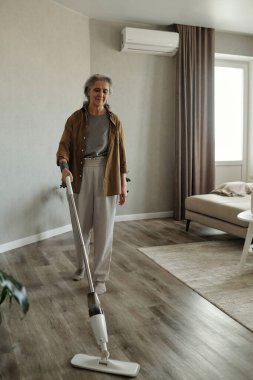 Modern oturma odasında yerleri temizleyen yaşlı bir kadın elektrikli süpürge kullanıyor, günlük giysiler giyiyor ve özenle çalışırken hafifçe gülümsüyor.