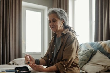 Yaşlı bir kadın, dizüstü bilgisayarda yazarken kanepede oturuyor, sakin bir ifadeyle ve kapalı mekan ışığıyla yumuşak gölgeler bırakıyor. Oturma odası ayarları sıcak ve tarafsız tonlarda.