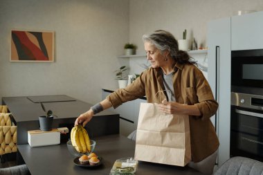 Çağdaş dekorasyon ve doğal ışıkla modern mutfakta paketleri açan yaşlı bir kadın. Mutfak adasında dikkatle hazırlanmış eşyalar.