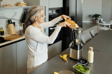 Blender 'da ıspanak, muz ve sütle karıştıran yaşlı bir kadın. Arka planda paslanmaz çelik aletler ve bitkilerle modern mutfakta duruyor.