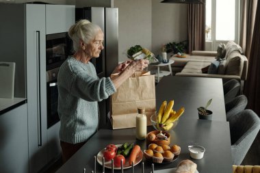 Gri saçlı yaşlı bir kadın modern bir mutfakta meyve, sebze ve şişelenmiş ürünler de dahil olmak üzere çeşitli yiyecek malzemeleriyle paketlerini açıyor.
