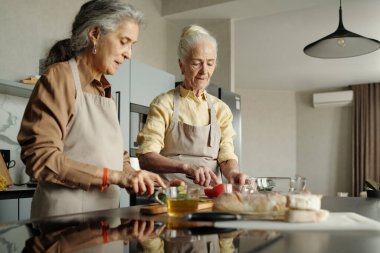Yaşlı adam ve kadın modern mutfakta yemek hazırlıyorlar, dikkatle, tezgahta sebze ve ekmek kesiyorlar ve önlük giyiyorlar.