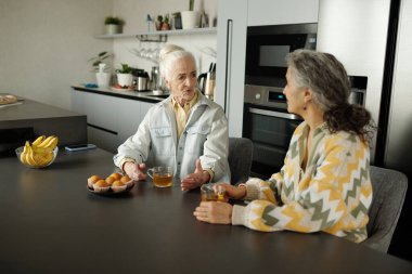 İki yaşlı kadın mutfak tezgahında oturmuş içki ve yumurtalarla sohbet ediyorlar. Modern mutfak aletleri ve dekorasyon arka planda görünür.