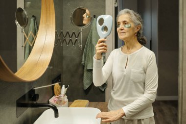 Banyodaki aynanın önünde duran yaşlı kadın el aynası ile cilt kremi sürerken yansımaya odaklanıyor.