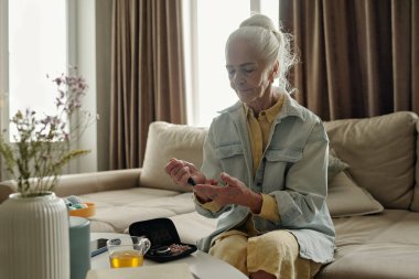 Kanepede oturan, glukometre kullanarak kan şekerini ölçen, etrafı çiçeklerle çevrili ve rahat oturma odasındaki masada çay içen yaşlı bir kadın. Sakin ve nişanlı görünüyor.