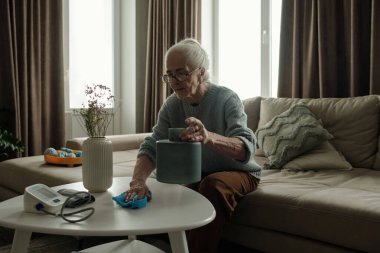 Modern ekipman ve teknolojiyi kullanarak rahat bir oturma odasında masayı temizleyen yaşlı bir kadın. Rahat bir kanepede oturuyor ve pencereden içeri ışık giriyor.