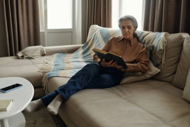 Oturma odasında rahat bir kanepede oturmuş kitap okuyan son sınıf öğrencisi bir kadın. 